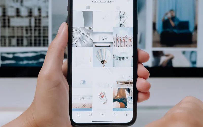 Manos sosteniendo un teléfono inteligente con la pantalla mostrando un perfil de Instagram con una variedad de publicaciones de diseño y arquitectura, frente a una computadora portátil con teclado y una persona desenfocada en el fondo
